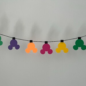 Puede incluir: Una guirnalda colorida con formas de cabeza de Mickey Mouse en varios colores, incluyendo amarillo, verde, morado, naranja y rosa. Las formas est&aacute;n ensartadas en una cuerda negra, creando una decoraci&oacute;n festiva y alegre.