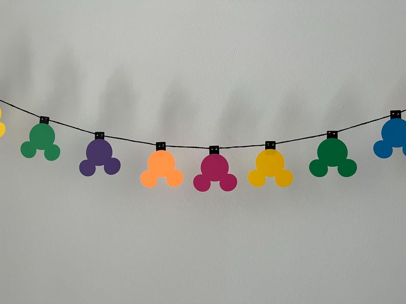 Puede incluir: Una guirnalda colorida con formas de cabeza de Mickey Mouse en varios colores, incluyendo amarillo, verde, morado, naranja y rosa. Las formas est&aacute;n ensartadas en una cuerda negra, creando una decoraci&oacute;n festiva y alegre.