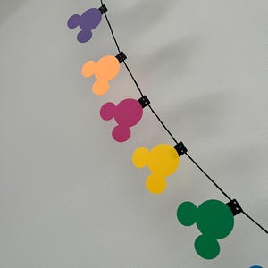 Puede incluir: Una colorida guirnalda con formas de cabeza de Mickey Mouse en verde, morado, naranja, rosa, amarillo y azul. Las formas est&aacute;n ensartadas en un cord&oacute;n negro, ideal para decoraci&oacute;n.