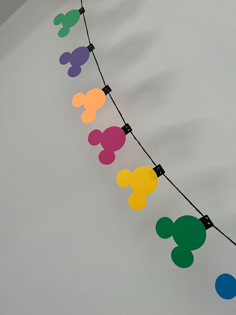 Puede incluir: Una colorida guirnalda con formas de cabeza de Mickey Mouse en verde, morado, naranja, rosa, amarillo y azul. Las formas est&aacute;n ensartadas en un cord&oacute;n negro, ideal para decoraci&oacute;n.