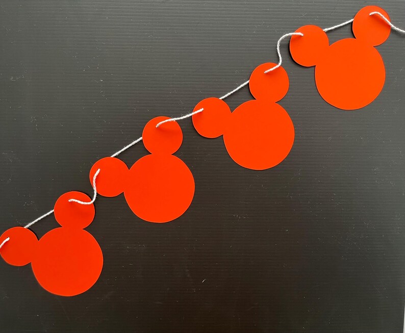 May include: A red and white string banner with four Mickey Mouse head cutouts. The banner is strung together with white string.