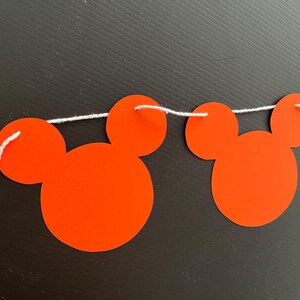 May include: A red and white Mickey Mouse themed banner with red paper cutouts of Mickey Mouse heads strung together with white string.