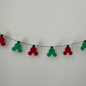 Puede incluir: Una guirnalda de recortes rojos y verdes con forma de Mickey Mouse, ensartados en un alambre negro. Los recortes est&aacute;n espaciados uniformemente y dispuestos sobre un fondo blanco liso. Una decoraci&oacute;n festiva.