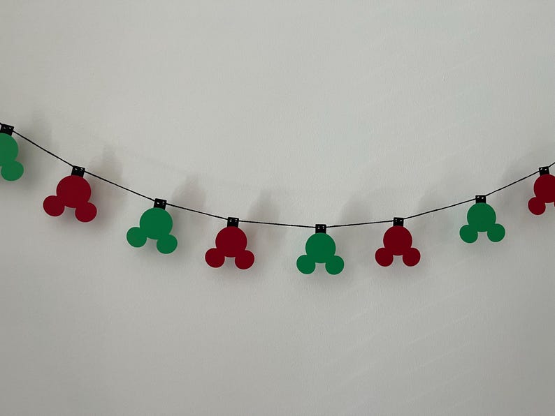 Puede incluir: Una guirnalda de recortes rojos y verdes con forma de Mickey Mouse, ensartados en un alambre negro. Los recortes est&aacute;n espaciados uniformemente y dispuestos sobre un fondo blanco liso. Una decoraci&oacute;n festiva.