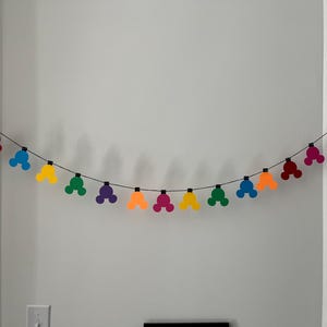 Puede incluir: Una colorida guirnalda con formas de cabeza de Mickey Mouse en rojo, azul, amarillo, verde, morado y naranja. La guirnalda est&aacute; ensartada en un alambre negro y cuelga contra una pared blanca.