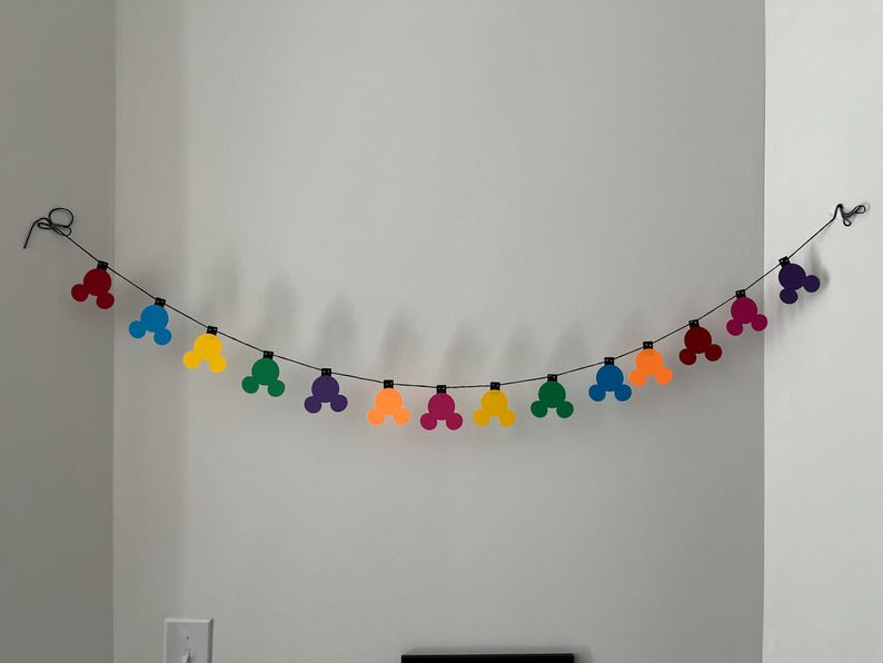 Puede incluir: Una colorida guirnalda con formas de cabeza de Mickey Mouse en rojo, azul, amarillo, verde, morado y naranja. La guirnalda est&aacute; ensartada en un alambre negro y cuelga contra una pared blanca.