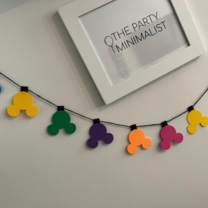 Puede incluir: Una colorida guirnalda con formas de cabeza de Mickey Mouse en azul, amarillo, verde, morado, naranja y rosa. La guirnalda est&aacute; ensartada en un cord&oacute;n negro y se muestra contra una pared blanca. Una impresi&oacute;n enmarcada con el texto "THE PARTY MINIMALIST" est&aacute; en el fondo.