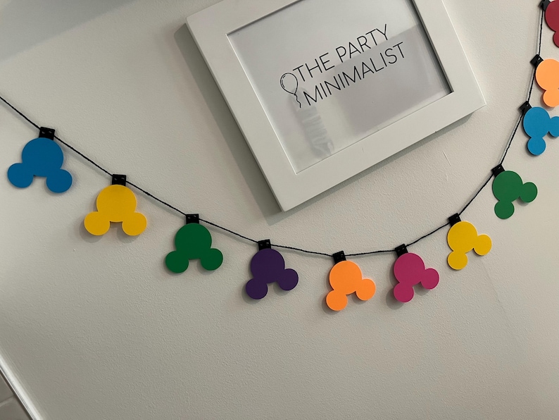 Puede incluir: Una colorida guirnalda con formas de cabeza de Mickey Mouse en azul, amarillo, verde, morado, naranja y rosa. La guirnalda est&aacute; ensartada en un cord&oacute;n negro y se muestra contra una pared blanca. Una impresi&oacute;n enmarcada con el texto "THE PARTY MINIMALIST" est&aacute; en el fondo.