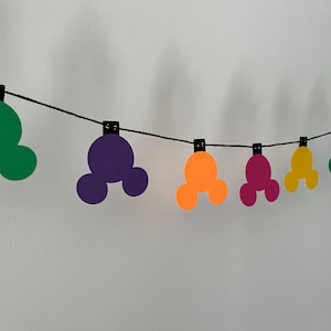 Puede incluir: Una guirnalda colorida con formas de cabeza de Mickey Mouse en verde, morado, naranja, rosa, amarillo y azul. Las formas est&aacute;n unidas a una cuerda negra con peque&ntilde;as pinzas negras, ideal para decorar.