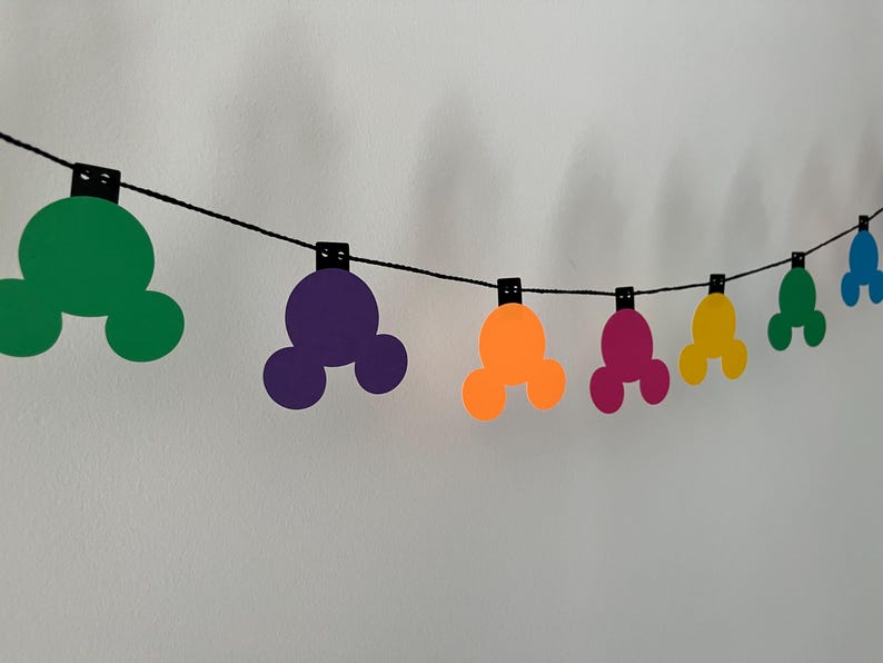 Puede incluir: Una guirnalda colorida con formas de cabeza de Mickey Mouse en verde, morado, naranja, rosa, amarillo y azul. Las formas est&aacute;n unidas a una cuerda negra con peque&ntilde;as pinzas negras, ideal para decorar.