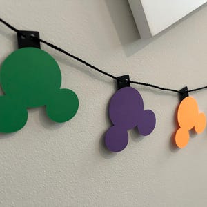 Puede incluir: Una guirnalda colorida con formas de cabeza de Mickey Mouse en verde, morado, naranja, rosa y amarillo. Las formas est&aacute;n ensartadas en un cord&oacute;n negro y sujetas con clips negros, creando una decoraci&oacute;n festiva.