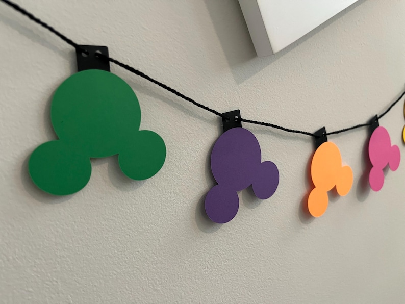 Puede incluir: Una guirnalda colorida con formas de cabeza de Mickey Mouse en verde, morado, naranja, rosa y amarillo. Las formas est&aacute;n ensartadas en un cord&oacute;n negro y sujetas con clips negros, creando una decoraci&oacute;n festiva.
