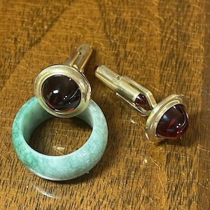 May include: A gold-toned ring with a large, round, dark red stone, a light green jade ring, and a pair of gold-toned cufflinks with matching dark red stones. The jewelry is displayed on a wooden surface.
