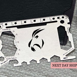 Könnte beinhalten: Eine silberne Multi-Tool-Karte mit einem Lineal in cm, einem Winkelmesser und einem Flaschenöffner. Sie hat ein schwarzes Grafikdesign und den Text "CM" und "Protractor 30".