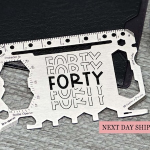 May include: A silver-colored stainless steel multi-tool card featuring a metric ruler along the top edge, a protractor, a bottle opener, a box opener, a wire cutter, and a nail puller. The center is engraved with the word FORTY in a bold font.