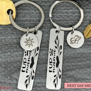 May include: Two silver keychains with engraved tags that read "Fur mama" and feature a small heart and a leaf design. One keychain has a small sun charm and the other has a small infinity symbol charm.