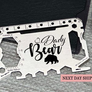Puede incluir: Una tarjeta multiherramienta plateada con las palabras "Dady Bear" y una silueta de oso. Incluye una regla con medidas en cm, un abrebotellas y un cortador de alambre. La tarjeta está sobre una superficie oscura.