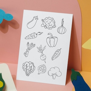 May include: A white card with black line drawings of various vegetables, including carrots, onions, eggplant, broccoli, and peppers. The card is on a pink surface with a blue cloud, a yellow star, and a green pea pod.