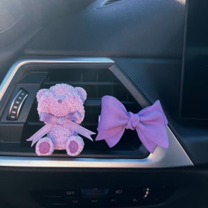 Könnte beinhalten: Ein lila Plüschteddybär und ein lila Schleifen-Autoluftaufhänger, der an der Lüftung eines Autos hängt.