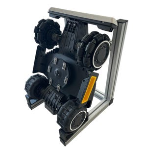 May include: A black and gray robotic chassis with multiple omni-directional wheels. The robot has a rectangular frame with a silver top and a yellow accent. The wheels are black with a unique tread pattern.
