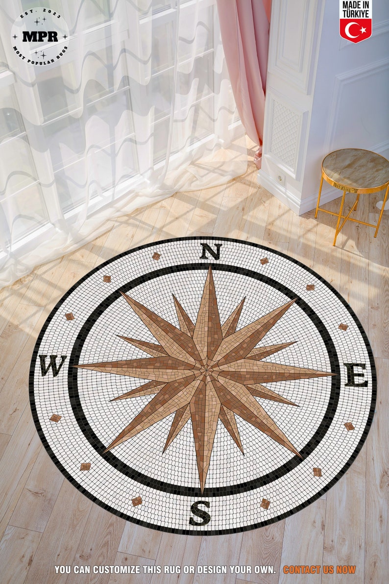 May include: A round, white and brown mosaic rug with a compass design. The rug features a black border with the letters N, S, E, and W. The rug is on a wooden floor.