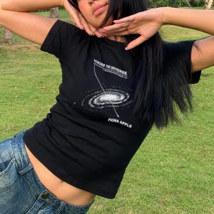 May include: A black short-sleeved t-shirt featuring a white graphic of a spiral galaxy. The text ACROSS THE UNIVERSE is printed above the galaxy, and the text FIONA APPLE is printed below it, with a thin white line connecting the two phrases.