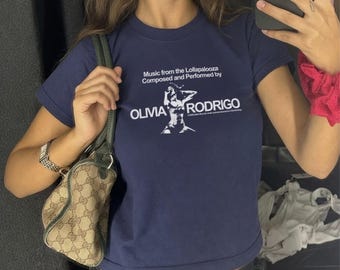 Op de afbeelding: Een marineblauw T-shirt met korte mouwen en witte tekst die luidt Music from the Lollapalooza Composed and Performed by OLIVIA RODRIGO, vergezeld van een witte grafische illustratie van een poserend persoon op het midden van de borst.