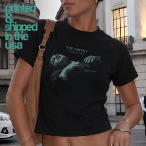 May include: A black short-sleeve t-shirt featuring a green graphic print of a person lying down with the text THE SMITHS The Queen Is Dead. The text Printed & Shipped in the USA is visible in the upper left corner of the image.