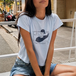 May include: A light blue short-sleeve t-shirt featuring a dark graphic print of a cartoon figure sitting on a dark shape. The text includes NARA, GIRLS, 1986, and Russian characters arranged in a circular pattern around the central illustration.