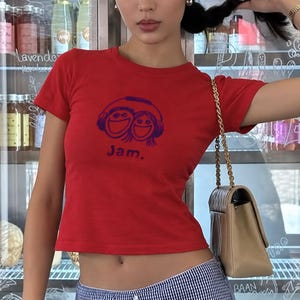 Pode incluir: Uma t-shirt vermelha de manga curta com uma estampa gráfica azul-escura de dois rostos sorridentes a usar auscultadores, com o texto "Jam." impresso logo abaixo da ilustração. A t-shirt tem um decote redondo e um corte curto e justo.