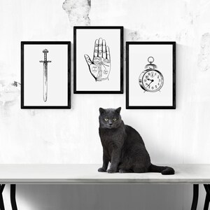 Peut inclure: Trois impressions encadr&eacute;es en noir et blanc : une &eacute;p&eacute;e, une main de chiromancie et un r&eacute;veil. Un chat gris est assis sur une surface blanche devant les impressions.