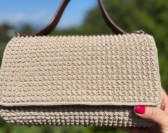 May include: A beige crochet handbag with a brown leather handle. The bag has a rectangular shape with a flap closure. The background is a blurred view of green foliage and a blue sky.