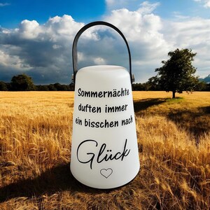Può includere: Una lanterna bianca con manico nero e la scritta "Sommernächte duften immer ein bisschen nach Glück" in nero. Un simbolo a forma di cuore è sotto la parola "Glück". La lanterna è posta in un campo di grano sotto un cielo blu.