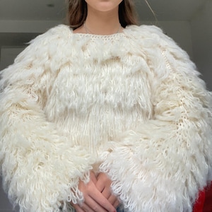 Puede incluir: Un suéter blanco peludo con mangas largas y cuello redondo. El suéter tiene una textura peluda y esponjosa.