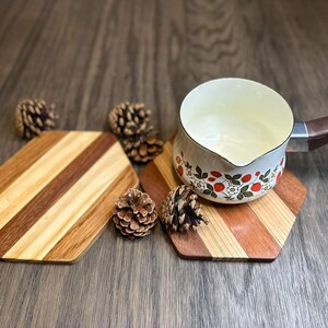 May include: Two hexagonal wooden trivets with a white enamel saucepan with a brown handle and a strawberry pattern on the side. The saucepan is sitting on one of the trivets. There are pine cones scattered around the trivets.