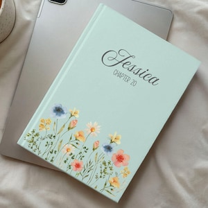 May include: A light blue journal with the name "Jessica" and "Chapter 20" in script. The bottom of the journal features a floral design with pink, yellow, and blue flowers. The journal is resting on a silver tablet.