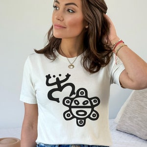 May include: White t-shirt with a black graphic of a stylized frog and a sun with a face. The sun has a circle around it with a series of smaller circles around the outer edge.