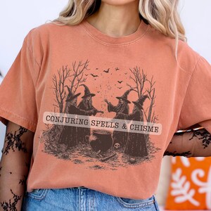 May include: Orange short-sleeved t-shirt with a graphic of four witches conjuring spells. The text "CONJURING SPELLS & CHISME" is printed across the image. The witches are in front of a bare tree and a cauldron.
