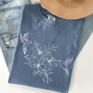 May include: A blue t-shirt with a white hummingbird and floral design. The shirt has the text "Comfort Colors" on the tag.