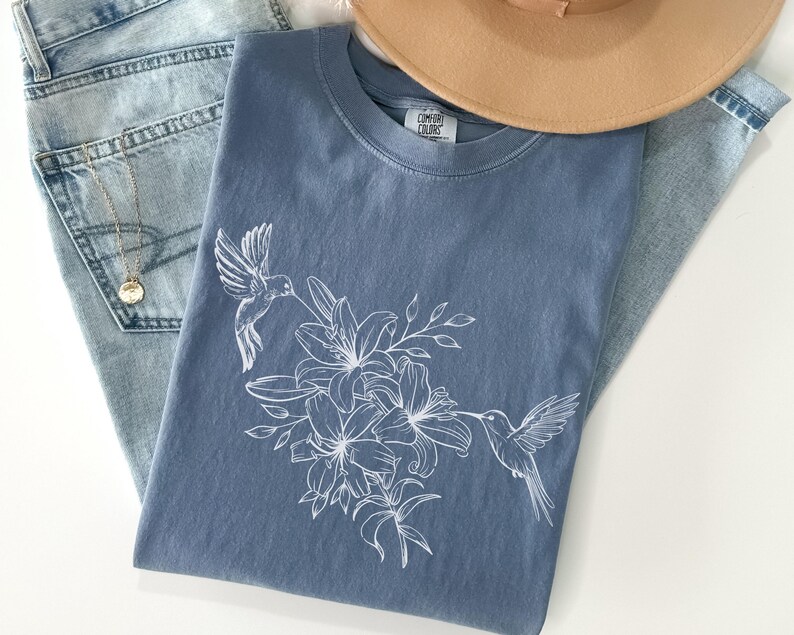 May include: A blue t-shirt with a white hummingbird and floral design. The shirt has the text "Comfort Colors" on the tag.