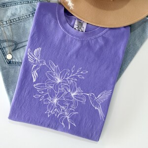 May include: A purple t-shirt with a white hummingbird and floral design. The design features two hummingbirds flying near a cluster of lilies.