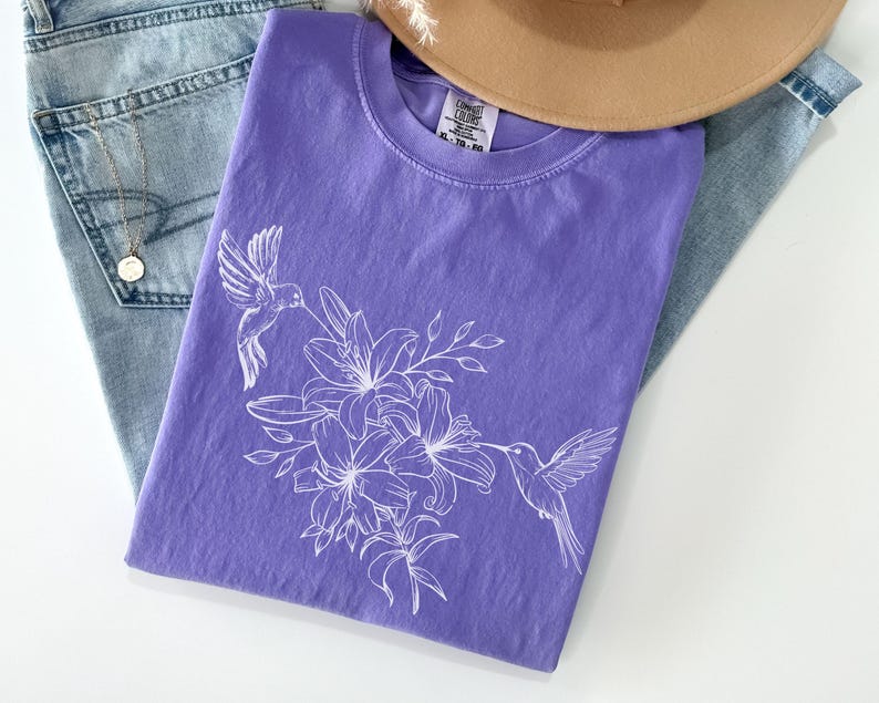 May include: A purple t-shirt with a white hummingbird and floral design. The design features two hummingbirds flying near a cluster of lilies.