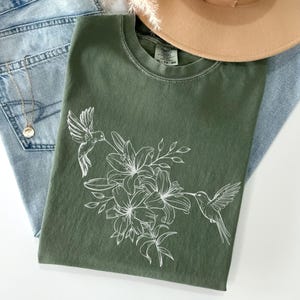May include: A green t-shirt with a white floral design and two hummingbirds. The design features lilies and leaves.