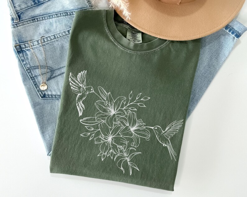 May include: A green t-shirt with a white floral design and two hummingbirds. The design features lilies and leaves.
