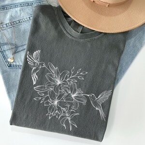 May include: A dark gray t-shirt with a white outline of hummingbirds and flowers. The shirt is folded and laid on top of blue jeans and a brown hat.