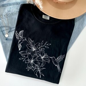 May include: Black t-shirt with a white hummingbird and floral design. The design features a hummingbird with its beak in a flower, surrounded by lilies and leaves.