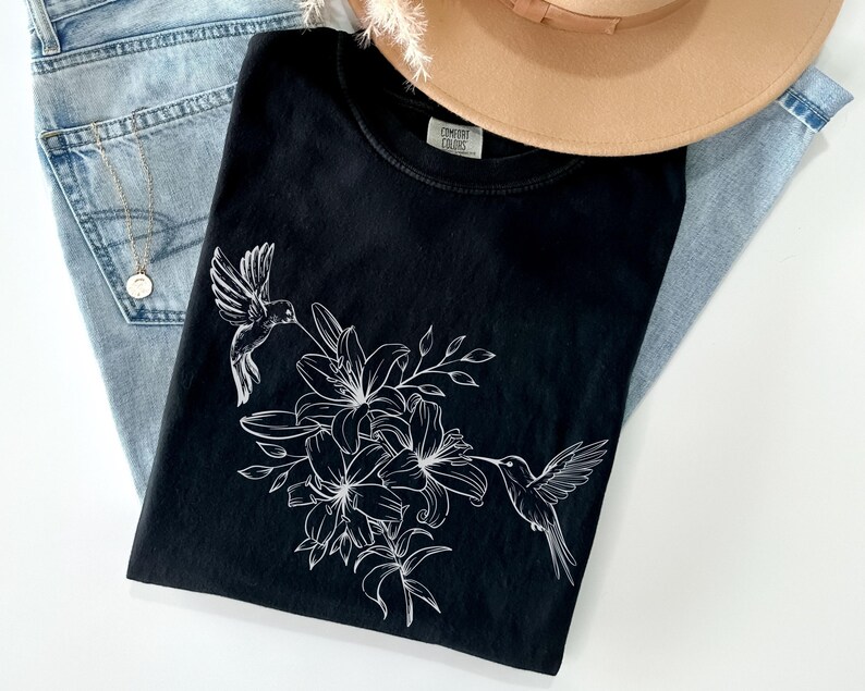 May include: Black t-shirt with a white hummingbird and floral design. The design features a hummingbird with its beak in a flower, surrounded by lilies and leaves.