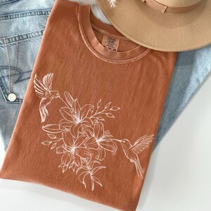 May include: A rust-colored t-shirt with a white hummingbird and lily flower design. The shirt has a crew neck and short sleeves.