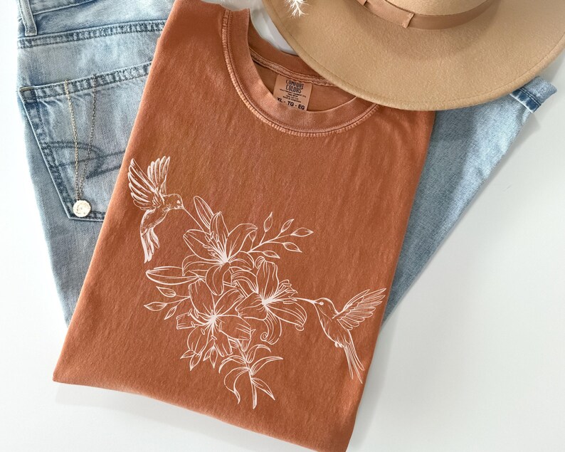 May include: A rust-colored t-shirt with a white hummingbird and lily flower design. The shirt has a crew neck and short sleeves.