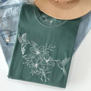 May include: A green t-shirt with a white hummingbird and floral design. The design features a hummingbird flying near a cluster of lilies.